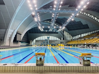 An indoor swimming pool with multiple lanes, brightly lit by overhead ceiling lights. The pool area is surrounded by large, arched structures, and an array of seats is visible on the right side. The water in the pool is clear, reflecting the bright lights above.