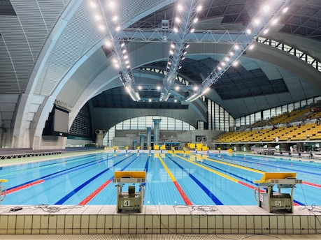 An indoor swimming pool with multiple lanes, brightly lit by overhead ceiling lights. The pool area is surrounded by large, arched structures, and an array of seats is visible on the right side. The water in the pool is clear, reflecting the bright lights above.