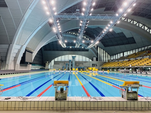 An indoor swimming pool with multiple lanes, brightly lit by overhead ceiling lights. The pool area is surrounded by large, arched structures, and an array of seats is visible on the right side. The water in the pool is clear, reflecting the bright lights above.
