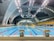 An indoor swimming pool with multiple lanes, brightly lit by overhead ceiling lights. The pool area is surrounded by large, arched structures, and an array of seats is visible on the right side. The water in the pool is clear, reflecting the bright lights above.