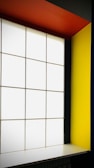 A modern interior featuring a large, grid-patterned frosted glass window flanked by bold, colorful walls. The right wall is a vivid yellow, and the ceiling is a striking orange. The overall design is minimalistic with high contrast colors.
