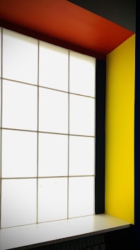 A modern interior featuring a large, grid-patterned frosted glass window flanked by bold, colorful walls. The right wall is a vivid yellow, and the ceiling is a striking orange. The overall design is minimalistic with high contrast colors.