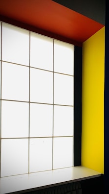 A modern interior featuring a large, grid-patterned frosted glass window flanked by bold, colorful walls. The right wall is a vivid yellow, and the ceiling is a striking orange. The overall design is minimalistic with high contrast colors.