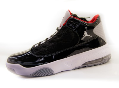 A high-top sneaker with a glossy black finish, featuring mesh panels and red accents on the interior lining. The shoe has a white midsole with a textured pattern and a translucent section on the heel. A distinctive logo is present on the side.