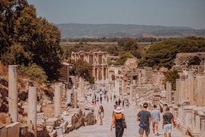 A historic archaeological site with ancient ruins, featuring a wide stone-paved pathway lined with columns and remnants of structures. Numerous tourists are walking along the pathway, some stopping to admire the ruins. In the background, a partially restored building with ornate details is visible. Lush greenery and hills frame the scenery.