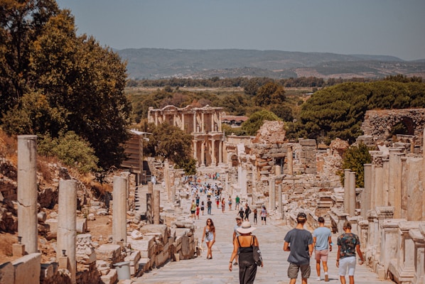 A historic archaeological site with ancient ruins, featuring a wide stone-paved pathway lined with columns and remnants of structures. Numerous tourists are walking along the pathway, some stopping to admire the ruins. In the background, a partially restored building with ornate details is visible. Lush greenery and hills frame the scenery.