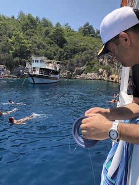 A man wearing a cap and holding a fishing reel stands on a boat overlooking a group of people swimming in clear blue water. A larger boat is anchored nearby against the backdrop of a lush, tree-covered hillside under a bright sky.