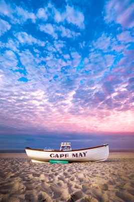 A boat with 'Cape May' written on it rests on a sandy beach as the sky above is filled with colorful clouds, ranging from blue to pink hues. The scene is calm and serene, with the vast beach extending to the horizon, bordered by the sea.