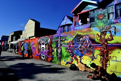 A vibrant street art mural covers the wall of a building, showcasing abstract and colorful designs with elements resembling futuristic figures and creatures. A person is seen working on the ground beside the mural, which features a variety of bold colors and imaginative shapes. Nearby buildings add an urban backdrop to the scene.