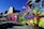 A vibrant street art mural covers the wall of a building, showcasing abstract and colorful designs with elements resembling futuristic figures and creatures. A person is seen working on the ground beside the mural, which features a variety of bold colors and imaginative shapes. Nearby buildings add an urban backdrop to the scene.