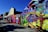 A vibrant street art mural covers the wall of a building, showcasing abstract and colorful designs with elements resembling futuristic figures and creatures. A person is seen working on the ground beside the mural, which features a variety of bold colors and imaginative shapes. Nearby buildings add an urban backdrop to the scene.
