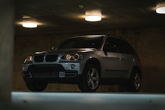 A sleek silver SUV is parked in a dimly lit underground parking garage. The vehicle features prominent front grilles and shiny metallic surfaces, surrounded by concrete walls and ceiling. Soft, circular ceiling lights cast a warm glow, creating shadows and highlights on the car’s exterior.