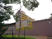 A multi-story building with a cross on top, suggesting it might be a school or religious institution. The structure features colorful accents with a pastel color scheme, including pink and yellow. A wall in the foreground has a poster with images of children, potentially students. Trees and greenery frame the scene, and the sky appears overcast.