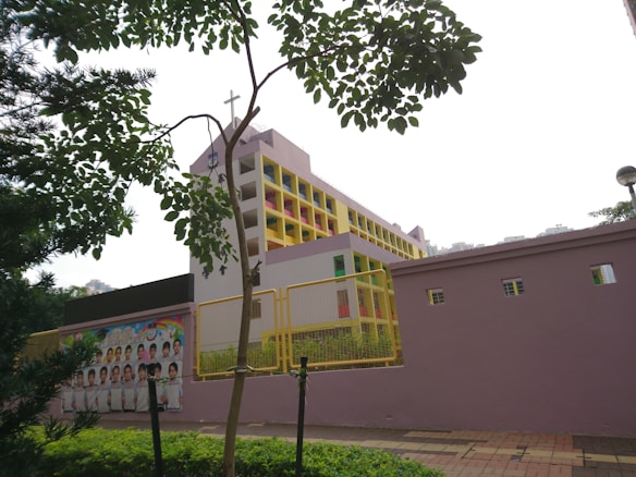 A multi-story building with a cross on top, suggesting it might be a school or religious institution. The structure features colorful accents with a pastel color scheme, including pink and yellow. A wall in the foreground has a poster with images of children, potentially students. Trees and greenery frame the scene, and the sky appears overcast.