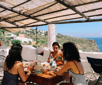 Three women are seated around a wooden table enjoying a meal under a shaded terrace. The table is filled with various foods and drinks. The setting overlooks a scenic view of hills, houses, and the distant ocean. The rustic roof structure and sunny weather contribute to a relaxed atmosphere.