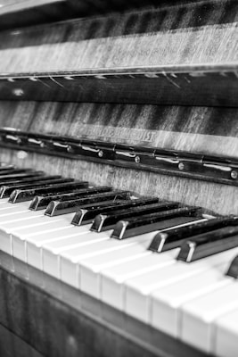A close-up view of a piano keyboard with keys arranged in a traditional layout. The image is in black and white, highlighting the contrast between the black and white keys. The wood grain of the piano's body is visible, adding texture to the composition.