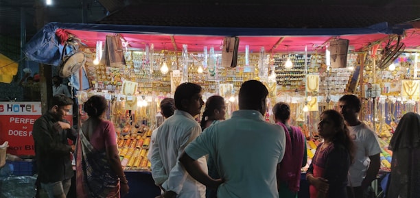 A bustling market stall at night, illuminated by hanging light bulbs, showcasing a wide array of jewelry and trinkets. Several people are gathered in front of the stall, engaged in browsing and shopping activities. The stall is richly decorated with necklaces, bangles, and other accessories hanging in an orderly fashion.