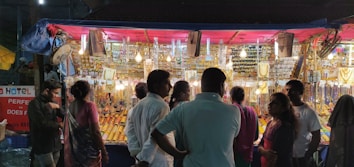 A bustling market stall at night, illuminated by hanging light bulbs, showcasing a wide array of jewelry and trinkets. Several people are gathered in front of the stall, engaged in browsing and shopping activities. The stall is richly decorated with necklaces, bangles, and other accessories hanging in an orderly fashion.