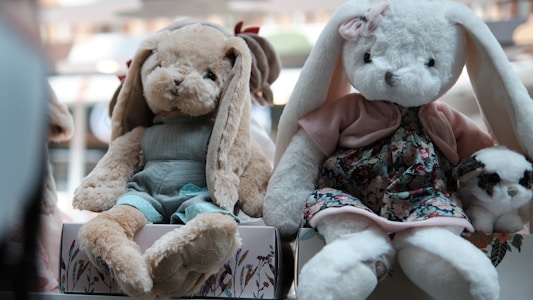 Two plush stuffed rabbits are dressed in detailed outfits. The rabbit on the left wears a teal outfit and has floppy light brown ears, while the rabbit on the right is in a floral dress with a pink cardigan and a matching pink bow on its ear. There is a small plush panda-like toy beside them. Both are sitting on decorative boxes and are displayed in a well-lit setting.