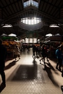 Interior view of a traditional market hall with a large skylight ceiling allowing sunlight to stream in, casting shadows on the tiled floor. Various stalls with colorful displays of fruits, vegetables, and other goods. People are walking around and shopping, some engaging with the vendors.