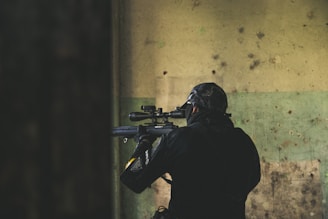 A person wearing tactical gear, including a helmet and face covering, is aiming a rifle with a scope. The individual is positioned indoors against a worn and textured wall with shades of green and beige.