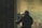 A person wearing tactical gear, including a helmet and face covering, is aiming a rifle with a scope. The individual is positioned indoors against a worn and textured wall with shades of green and beige.