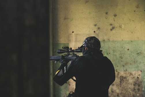 A person wearing tactical gear, including a helmet and face covering, is aiming a rifle with a scope. The individual is positioned indoors against a worn and textured wall with shades of green and beige.