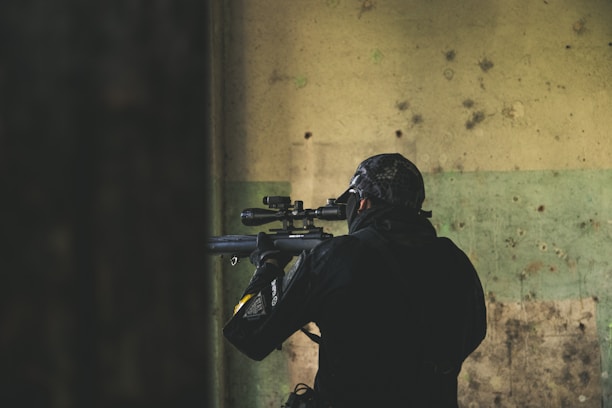 A person wearing tactical gear, including a helmet and face covering, is aiming a rifle with a scope. The individual is positioned indoors against a worn and textured wall with shades of green and beige.