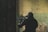 A person wearing tactical gear, including a helmet and face covering, is aiming a rifle with a scope. The individual is positioned indoors against a worn and textured wall with shades of green and beige.
