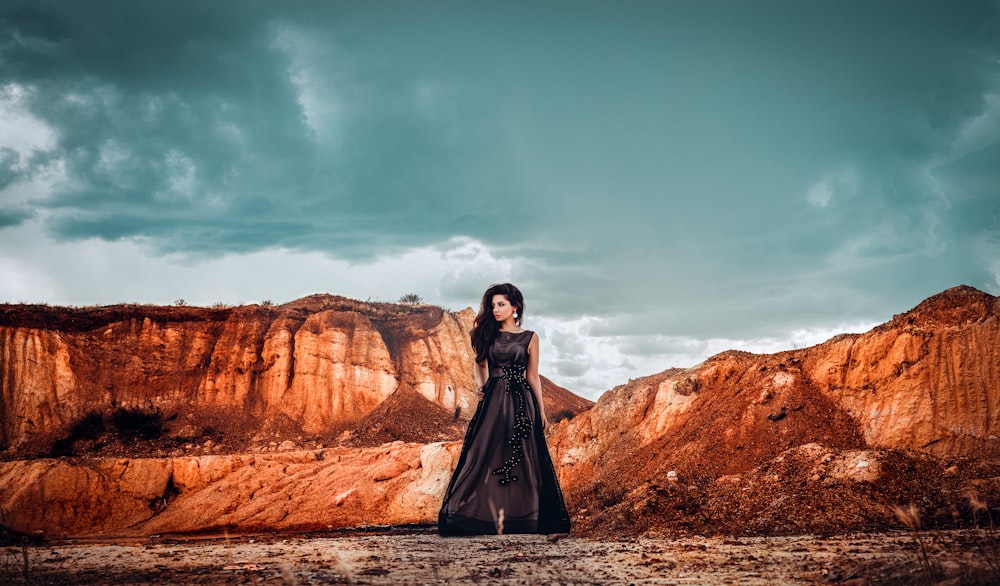 Femme en robe noire debout sur la formation rocheuse brune sous le ciel bleu pendant la journée