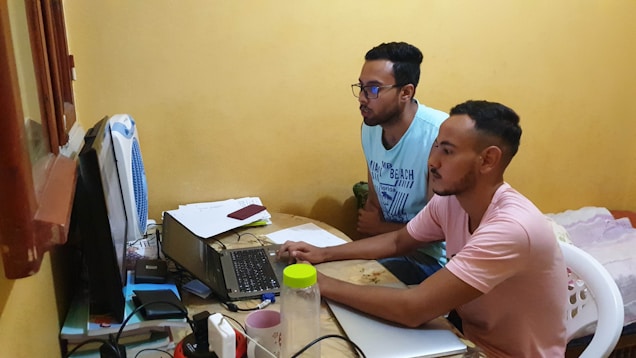 Two people are focused on a laptop screen in a small, cozy room. Papers and books are scattered on the desk, with a fan positioned at one end. They appear to be working or studying together, sitting on plastic chairs.