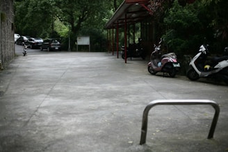 An outdoor parking area with several parked vehicles, including motorcycles and scooters, beside a building. Trees and greenery surround the space, and a few cars are seen parked in the distance. The ground is a smooth concrete surface, and an empty bicycle rack is in the foreground.