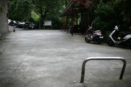 An outdoor parking area with several parked vehicles, including motorcycles and scooters, beside a building. Trees and greenery surround the space, and a few cars are seen parked in the distance. The ground is a smooth concrete surface, and an empty bicycle rack is in the foreground.