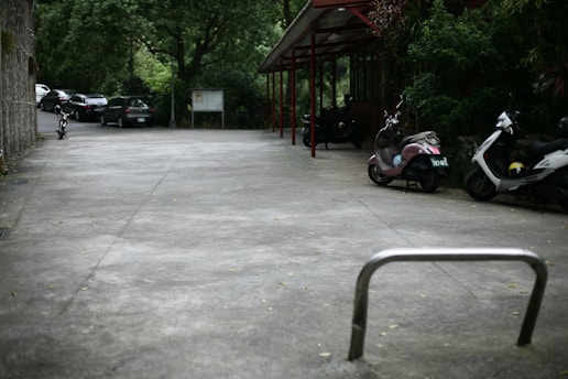 An outdoor parking area with several parked vehicles, including motorcycles and scooters, beside a building. Trees and greenery surround the space, and a few cars are seen parked in the distance. The ground is a smooth concrete surface, and an empty bicycle rack is in the foreground.
