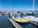 A marina filled with various boats and yachts docked alongside piers. A bright yellow inflatable boat is prominent in the foreground, tied to a wooden pier. Numerous sailboats with tall masts are lined up in the background under a clear blue sky.