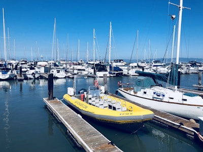A marina filled with various boats and yachts docked alongside piers. A bright yellow inflatable boat is prominent in the foreground, tied to a wooden pier. Numerous sailboats with tall masts are lined up in the background under a clear blue sky.