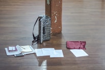An accordion is positioned upright against a wooden pillar on a wooden floor. In front of the accordion, there are various papers spread out, a small notebook, and what appears to be a mobile phone. A maroon-colored cloth is also present among the items on the floor.