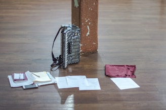 An accordion is positioned upright against a wooden pillar on a wooden floor. In front of the accordion, there are various papers spread out, a small notebook, and what appears to be a mobile phone. A maroon-colored cloth is also present among the items on the floor.