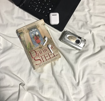 A book titled '44 Charles Street' by Danielle Steel rests on a bed covered with white sheets. Next to the book is a set of wireless earphones and a digital camera. Part of a laptop and a small, square electronic device are also visible.