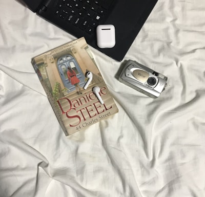 A book titled '44 Charles Street' by Danielle Steel rests on a bed covered with white sheets. Next to the book is a set of wireless earphones and a digital camera. Part of a laptop and a small, square electronic device are also visible.