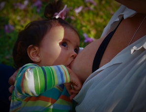 A young child is breastfeeding, with a focus on the child's face and the mother's breast. The child has dark hair and is wearing a colorful, striped shirt. The background is blurred, with hints of greenery and flowers.