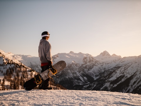 A person wearing winter clothing stands on a snowy slope holding a snowboard, with a vast mountain range in the background, bathed in warm sunlight.