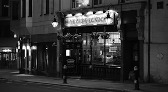 An old-fashioned pub with an illuminated sign sits quietly on a dimly lit street. Ornate street lamps and hanging flower baskets add to the quaint atmosphere, and the building features large windows and intricate architectural details.