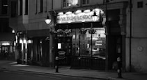 An old-fashioned pub with an illuminated sign sits quietly on a dimly lit street. Ornate street lamps and hanging flower baskets add to the quaint atmosphere, and the building features large windows and intricate architectural details.