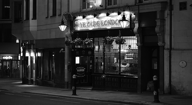 An old-fashioned pub with an illuminated sign sits quietly on a dimly lit street. Ornate street lamps and hanging flower baskets add to the quaint atmosphere, and the building features large windows and intricate architectural details.