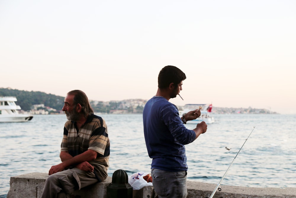 man in blue jacket sitting beside man in black jacket holding fishing rod during daytime