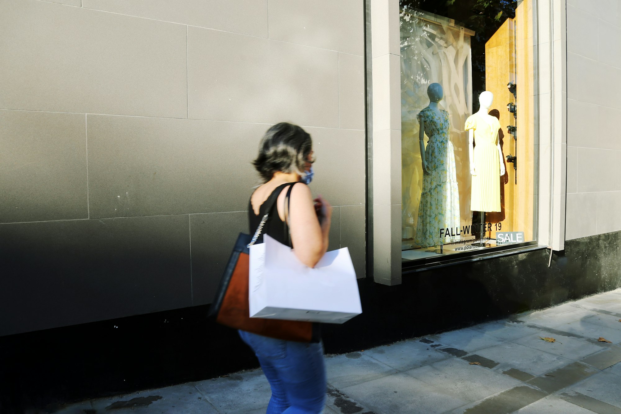 Mulher andando pela calçada com sacola de compras no ombro, em frente a uma vitrine