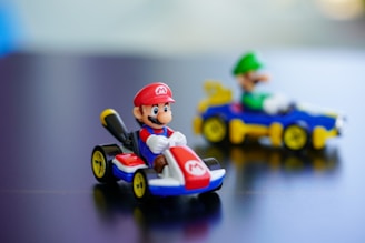 Two small toy cars with characters seated in them on a smooth surface. The closer toy features a figure in a red hat and blue outfit, while the second toy in the background has a figure in a green hat and shirt.
