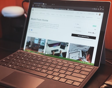 A laptop with a keyboard is open on a desk displaying a webpage about remote work. The screen shows images and text related to working from home, with tabs visible at the top of the browser. There is a smart speaker to the left and part of a chair visible at the edge of the frame.