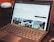 A laptop with a keyboard is open on a desk displaying a webpage about remote work. The screen shows images and text related to working from home, with tabs visible at the top of the browser. There is a smart speaker to the left and part of a chair visible at the edge of the frame.